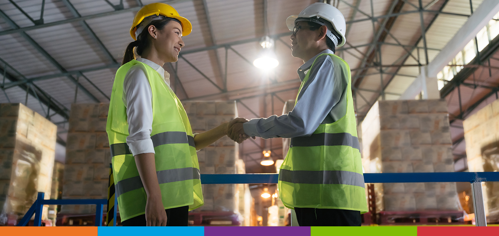 A woman in a yellow hard hat shaking hands with a man in a white hard hat in a factory full of pallets of boxes